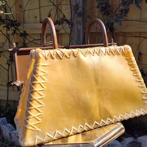 May include: A light brown leather handbag with a unique design. The bag has a woven leather strap and a decorative stitch pattern.