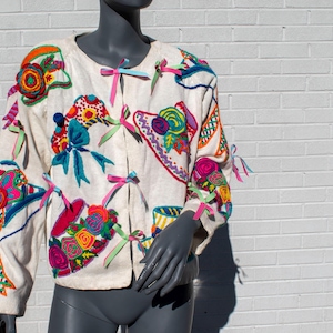 May include: An off-white long-sleeve jacket featuring colorful embroidered floral and hat motifs. The front closure is secured with multiple thin, multi-colored ribbon ties in shades of pink, blue, and green, adding texture to the garment.