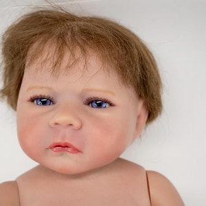 May include: A lifelike baby doll with a fair complexion, blue eyes, and light brown hair. The doll's face has a realistic appearance, with detailed features and a slightly pursed mouth. The doll's skin has a subtle blush, and the hair is styled in a soft, natural way.
