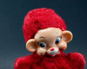May include: A vintage toy monkey with a red fuzzy hat and body. The monkey has a cream-colored face with blue eyes, a red nose, and large ears. The toy is a close-up shot against a dark background.