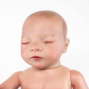May include: A lifelike baby doll with closed eyes, peach skin, and subtle red hair. The doll's face has realistic features, including a small nose and slightly parted lips. The doll is designed to resemble a newborn.