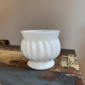 May include: A small, white milk glass bowl featuring a rounded body with a raised, vertical leaf-like pattern around the exterior. The bowl has a smooth, flared rim and sits on a circular pedestal base, creating a classic, textured appearance.