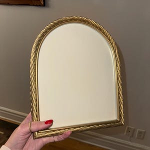 May include: A gold-coloured, arched picture frame with a cream-coloured interior. The frame has a rope-like texture around the edges. The frame is held by a person with red nail polish.