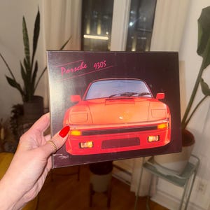 May include: A rectangular black box featuring a graphic illustration of a bright orange Porsche 930S sports car on the front. The text Porsche 930S is printed in a stylized pink script in the upper left corner of the box cover.