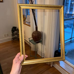 以下が含まれることがあります： ロープのような詳細なボーダーが付いた、長方形の金色のフォトフレーム。窓の前にフレームが保持され、その装飾的なデザインと温かみのあるメタリックカラーが示されています。フレームは空で、写真やアートワークの準備ができています。
