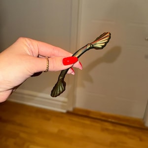 Può includere: Una maniglia per mobili in stile antico con finitura bronzo. La maniglia presenta un design curvo con estremità decorative a forma di conchiglia. L'hardware è per mobili.