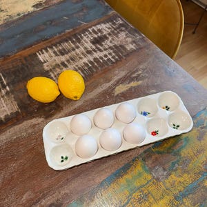 May include: A white ceramic egg holder with eight eggs and four decorative egg cups, each with a floral design. Two yellow lemons sit beside the egg holder on a rustic wooden table.