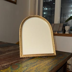 May include: A gold-coloured, arched picture frame with a cream-coloured interior. The frame has a rope-like texture around the edges. The frame is on a multi-coloured wooden table.