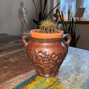 May include: A small, round, spiky cactus with yellow needles sits inside a plain terracotta pot, which is placed within a larger, decorative, two-handled ceramic planter featuring a glossy, mottled reddish-brown glaze and embossed scrollwork.