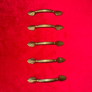 May include: Five antique brass drawer pulls with a shell-like design on a vibrant red background. Each pull features a curved handle with horizontal lines, adding a touch of vintage elegance. Ideal for furniture restoration or adding a classic touch to cabinets.