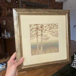 May include: A square framed wall art piece featuring a print of two birch trees with brown foliage against a soft cream and blue background. The artwork is enclosed in a textured, metallic gold-toned frame with a wide cream-colored matting border.