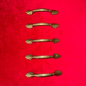 May include: Five antique brass drawer pulls with a shell-like design against a vivid red backdrop. Each pull has a curved handle with horizontal lines, offering a vintage aesthetic. Suitable for furniture restoration or adding a classic touch to cabinets.