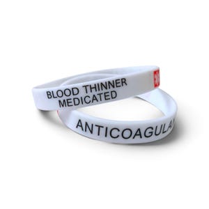 May include: Two white silicone medical alert wristbands stacked on each other. The top band features the text BLOOD THINNER MEDICATED in black ink and a red medical star symbol. The bottom band displays the text ANTICOAGULANT in black ink.