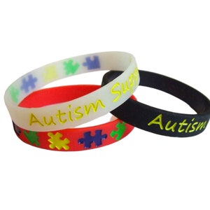 Op de afbeelding: Drie rubberen armbanden in wit, rood en zwart. De witte armband heeft kleurrijke puzzelstukjes en de woorden "Autism Support" in geel. De rode armband heeft puzzelstukjes. De zwarte armband heeft het woord "Autism" in geel.
