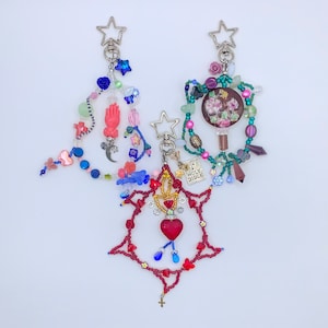 May include: Two handmade keychains with star-shaped clasps. One features a red heart pendant within a red beaded star, and the other has a pink hand charm. Both have colorful beads and charms, including crosses, moons, and flowers.