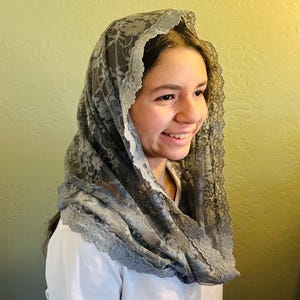 May include: A gray lace veil with a scalloped edge. The veil is draped over the head and shoulders. The lace pattern is floral. The person is wearing a white t-shirt.