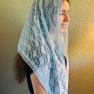 Puede incluir: Un velo de encaje azul claro con un patrón floral. El velo está drapeado sobre la cabeza y los hombros. Los bordes están acabados con un ribete festoneado. La persona lleva una camiseta negra.