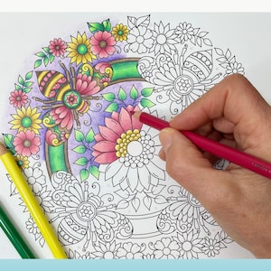 May include: A close-up of a hand coloring a detailed mandala design with colored pencils. The mandala features a bee, flowers, and leaves. The design is partially colored in shades of pink, yellow, and green.