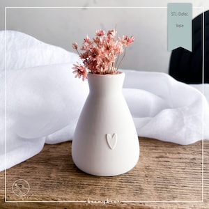 Könnte beinhalten: Eine kleine, weiße Keramikvase mit herzförmigem Design, die einen Strauß getrockneter rosa Blumen enthält. Die Vase hat eine abgerundete Form und einen schmalen Hals. Der Text "STL-Datei: Vase" befindet sich oben rechts.