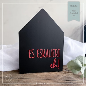 Könnte beinhalten: Ein schwarzes hausförmiges Schild mit dem roten Schriftzug "ES ESKALIERT eh!". Das Schild steht auf einer Holzoberfläche, mit einem weißen Tuch und Grünzeug im Hintergrund. Ein kleines türkisfarbenes Etikett oben rechts enthält Text.