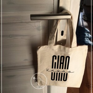 May include: A white canvas tote bag with black text that reads "Gian Kakao Vivo". The bag is hanging on a door handle.