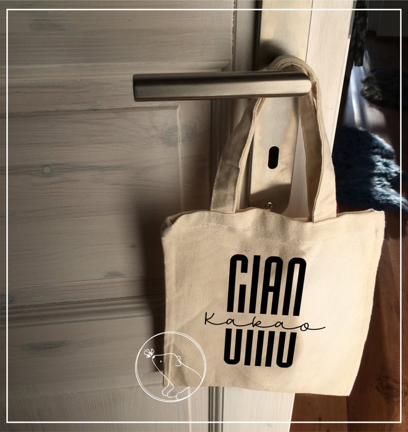 May include: A white canvas tote bag with black text that reads "Gian Kakao Vivo". The bag is hanging on a door handle.