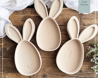 May include: Three beige Easter bunny-shaped trays with raised edges, each with a different design. One has a heart, another has a face, and the third is plain. The trays are on a wooden surface.