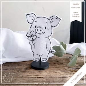 Peut inclure: Figurine de cochon en feutre blanc tenant un trèfle à quatre feuilles, monté sur un socle noir. Le cochon a un visage souriant et est souligné en noir. Le fond comprend un tissu blanc et une surface en bois.