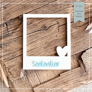 May include: A white square photo frame with a cutout heart shape in the bottom right corner and the light blue text Seelentier attached to the bottom border. The frame has a square opening in the center and a smooth, flat surface finish.