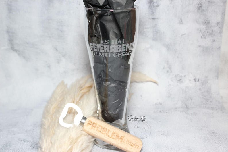 May include: A clear glass beer stein with the text "Es hat Feierabend zu mir gesagt" etched on the side. A wooden bottle opener with the text "Problem?  &Ouml;ffen!" is in the foreground.