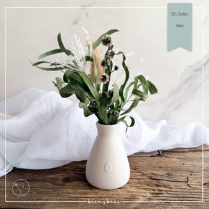 Könnte beinhalten: Eine kleine, weiße Keramikvase mit einem Trockenblumenarrangement. Das Arrangement umfasst grüne Blätter, weiße, federartige Akzente und beige Gräser. Die Vase hat eine abgerundete Form und ein kleines geprägtes Design. Der Text "STL-Datei: Vase" ist sichtbar.