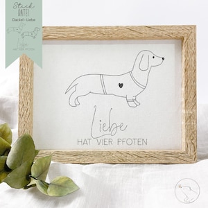 Pode incluir: Uma impressão emoldurada com uma ilustração em preto e branco de um dachshund vestindo um suéter com um coração. O texto "Liebe hat vier Pfoten" está impresso abaixo da ilustração.