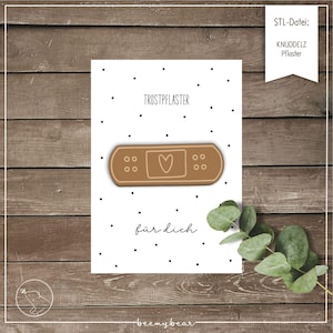 May include: A white card with black polka dots features a brown bandage with a heart design. The German text "Trostpflaster" and "für dich" are printed on the card. A sprig of green leaves is in the lower right corner.