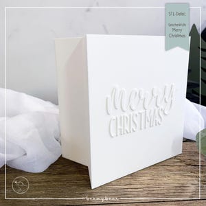May include: A white, folded paper gift box with the embossed words "Merry Christmas" on the front. The box is on a wooden surface, with a white fabric and a green accent in the background. The text "STL-Datei: Geschenkfülle Merry Christmas" is in the upper right corner.