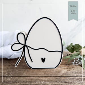 May include: A white Easter egg-shaped decoration with a black outline and a black bow on the side. A black heart is at the bottom. The text "STL-Datei: Ei mit Schleife" is in the upper right corner. The logo "beemybear" is at the bottom.