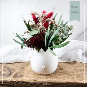 May include: A white, egg-shaped vase with a heart detail, filled with a vibrant arrangement of dried flowers in shades of red, green, and cream. The vase sits on a wooden surface, with text that reads "STL-Datei: Eierschale".