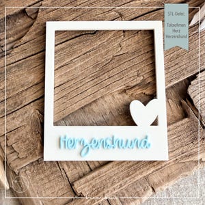 May include: A white rectangular photo frame with a square opening, featuring a white heart shape on the bottom right corner and the light blue script text "Herzenshund" embossed on the bottom border of the frame.