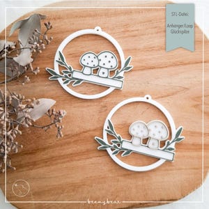 Op de afbeelding: Twee witte ornamenten met een paddenstoelthema, met een cirkelvormige lijst en een blanco banner. De ornamenten zijn versierd met groene bladeren en staan op een houten achtergrond. De tekst "STL-Datei: Anhänger/Loop Glückspilze" is zichtbaar.