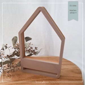 May include: A light brown wooden house-shaped frame with a shelf. The frame is on a wooden surface. The text "STL-Datei: Hauschen-rahmen +" is visible in the upper right corner of the image.