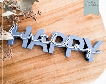 May include: A blue and white "Happy Birthday" cake topper. The word "Happy" is in blue block letters, with a white cursive "birthday" overlay. The topper is set on a wooden surface, with sprigs of dried flowers in the background.