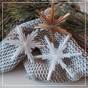 Könnte beinhalten: Drei Papierstern-Ornamente, zwei weiße und ein brauner, sind auf einem grauen Strickstoff angeordnet. Die weißen Sterne sind aus gefaltetem Papier gefertigt und haben ein zartes, filigranes Aussehen. Der braune Stern ist aus dickerem Papier gefertigt und hat ein rustikaleres Aussehen.