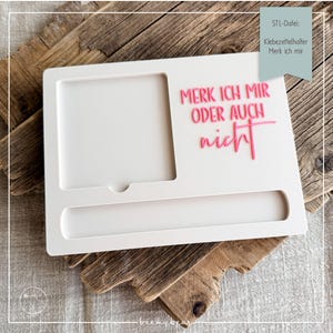 Peut inclure: Un organiseur de bureau rectangulaire blanc avec un cadre photo, une fente et le texte rose "MERK ICH MIR ODER AUCH nicht". L'organisateur est sur une surface en bois. Le texte "STL-Datei: Klebezettelhalter Merk ich mir" est également visible.