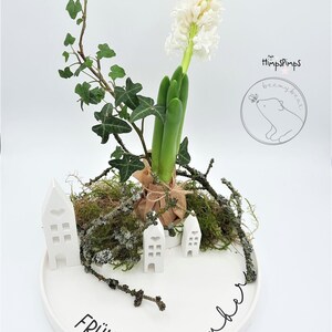 以下が含まれることがあります： 白いヒヤシンスの花の飾り付け。白い陶器の皿に、