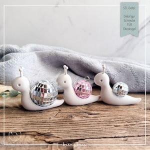 Op de afbeelding: Drie witte slakfiguren met gladde lichamen en voelsprieten. Elke slak heeft een reflecterende discobal als schelp. De linker slak heeft een grote zilveren discobal, de middelste een roze discobal en de rechter een kleine zilveren discobal.