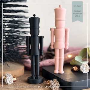 May include: Two nutcracker figurines, one in matte black and one in soft pink, standing upright. Each figure features a tall hat, cylindrical body, and articulated limbs. A small sign in the upper right corner displays the text STL-Datei: Dekofigur Nussknacker schlicht.