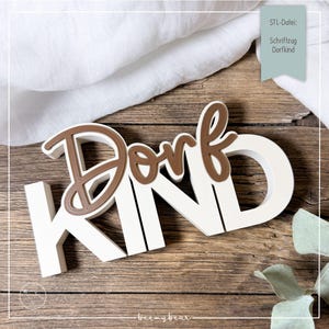 Könnte beinhalten: Ein Holzschild mit dem Wort "Dorfkind" in Braun und Weiß. Das Wort ist in einer stilisierten Schriftart gehalten. Das Schild befindet sich auf einer Holzoberfläche.