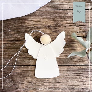 Può includere: Un ornamento a forma di angelo bianco con un dettaglio a cuore, una testa di perline di legno e un filo bianco per appenderlo. L'angelo ha ali dettagliate ed è posto su uno sfondo di legno rustico. Il testo "Engel" è visibile.