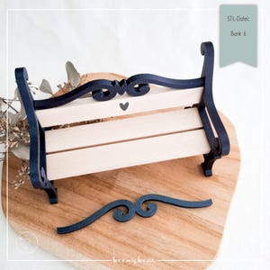Könnte beinhalten: Eine Miniatur-Holzbank mit marineblauem Rahmen und heller Holzbankfläche. Die Bank hat dekorative Schnörkel und ein kleines Herzdetail. Der Text "STL-Datei: Bank 6" ist sichtbar. Ein separates marineblaues Schnörkelstück ist ebenfalls vorhanden.