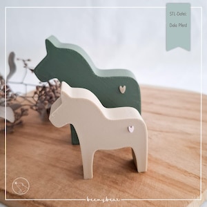 Può includere: Due cavalli di Dala in legno, uno verde salvia e uno beige, sono esposti su una superficie di legno. Ogni cavallo ha un piccolo dettaglio a forma di cuore. Il testo "STL-Datei: Dala Pferd" è visibile in alto a destra.