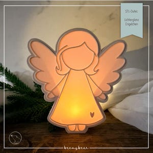 May include: A light-up angel figurine with a warm orange glow, featuring a simple outline of a figure with wings and a small heart on the dress. A light blue tag in the corner displays the text STL-Datei: Lichterglanz Engelchen.